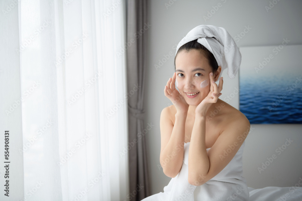 Happy young Asian woman applying face lotions while wearing a towel and touching her face. Daily makeup and skincare Happy young Asian woman applying face lotions while wearing a towel and touching her face. Daily makeup and skincare