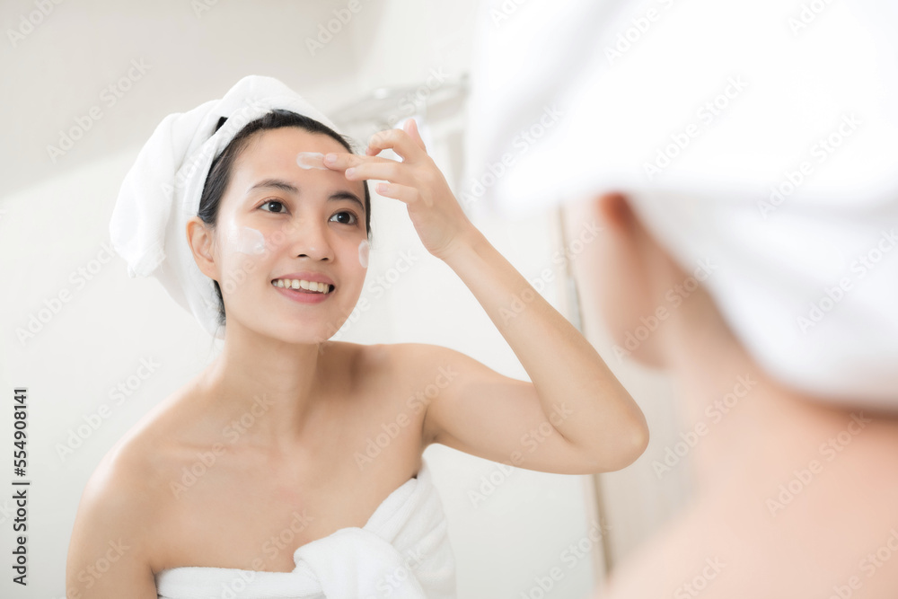 Happy young Asian woman applying face lotions while wearing a towel and touching her face in bathroom Happy young Asian woman applying face lotions while wearing a towel and touching her face in bathroom