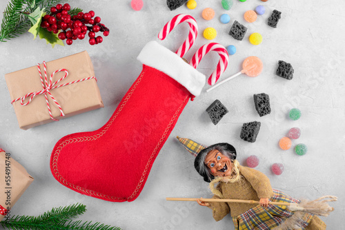 Foto The funny witch Befana and red stocking with sweet coal and candy on light gray background