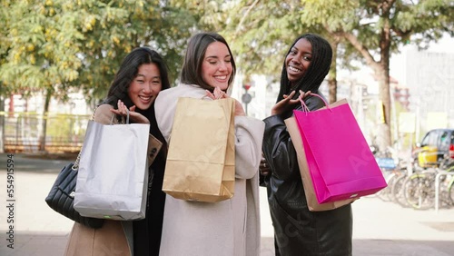 A group of young happy diverse women walking with shopping bags and turn back smiling to look and wave to the camera in a city urban street. Consumism Concept. High quality 4k slowmotion footage photo