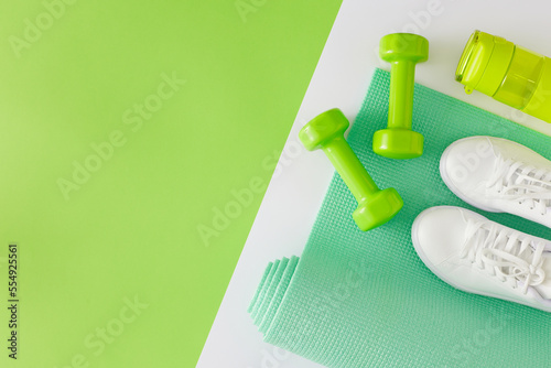 Fitness concept. Flat lay composition of dumbbells exercise mat bottle of water sports shoes on white and green background with copy space. Minimal athletics accessories idea. photo