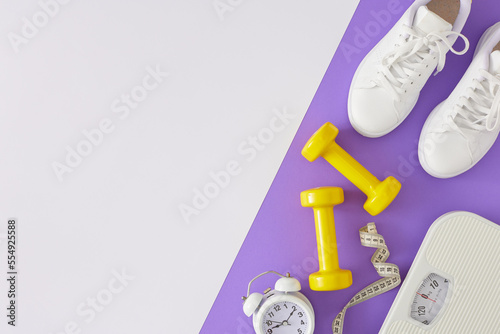 Fitness concept. Top view composition of yellow dumbbells sports shoes scales tape measure and alarm clock on white and violet background with copy space. Minimal sport idea.