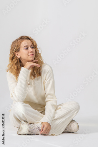 Pretty young blonde woman with curly hair is sitting on the floor on a white background. The girl is dressed in a warm white knitted suit.
