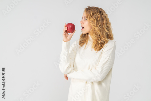A young pretty curly blonde woman holds a red apple in her hands. The girl looks happy and smiles, she is dressed in a white knitted suit. Near the model there is a lot of space for a