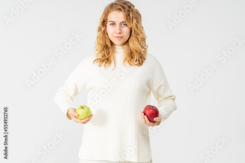 Young pretty curly blonde woman holds green and red apple in her hands. The girl looks happy and smiles, she is dressed in a white knitted suit. Near the model there is a lot of space for advertising
