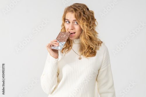 Young pretty curly blonde tries a chocolate bar with nuts. The girl looks happy and smiles, she is dressed in a white knitted suit. Model posing on white background