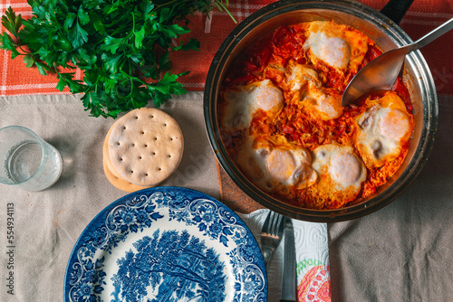 Top view of a cooking pan with shakshuka and a ladle, empty plate, fresh parsley and gluten free bread photo