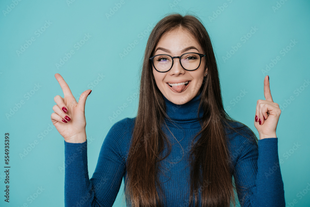 Pretty European woman in blue sweater at studio over turquoise backdrop in glasses, look at camera, points up with both hand by index fingers, shows tongue. Look up, mockup. Level up concept, fun. Pretty European woman in blue sweater at studio over turquoise backdrop in glasses, look at camera, points up with both hand by index fingers, shows tongue. Look up, mockup. Level up concept, fun.