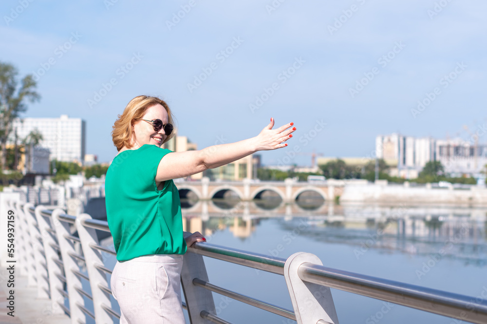 Portrait of a girl standing on the bank of the river holding on to the fence against the background of a blurred city.