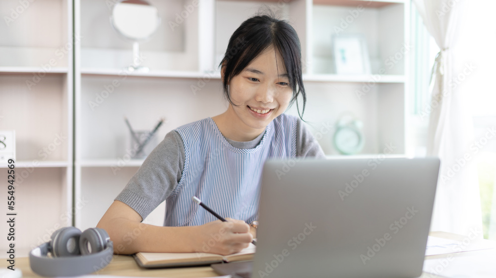 Asian woman taking notes in notebook while studying online in laptop at home, Video chat, Online communication , Stay home, New normal, Distance learning.., Social distancing, Learn online... Asian woman taking notes in notebook while studying online in laptop at home, Video chat, Online communication , Stay home, New normal, Distance learning.., Social distancing, Learn online...