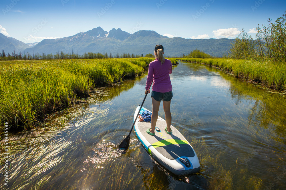 View from behind of a caucasian woman paddle boarding at Rabbit Slough, towards Twin Peaks in the Chugach Mountains, on a sunny, summer day with a blue sky in Wasilla; Alaska, United States of America View from behind of a caucasian woman paddle boarding at Rabbit Slough, towards Twin Peaks in the Chugach Mountains, on a sunny, summer day with a blue sky in Wasilla; Alaska, United States of America