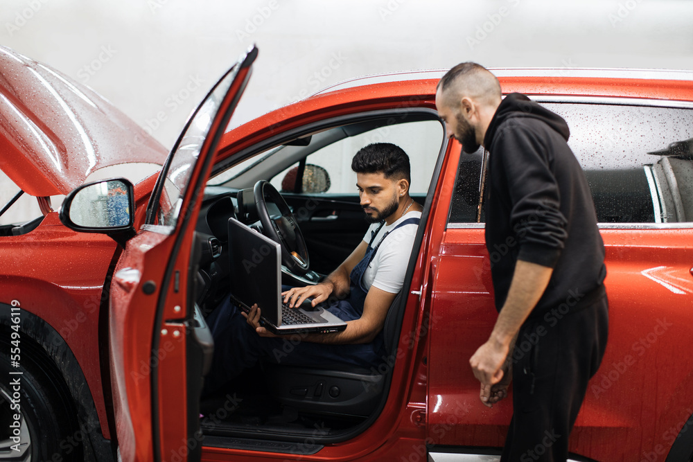 Two young male mechanic using laptop and tablet, recording automobile engine checking collecting detailed information during his work on car workshop. Service maintenance during engine repair.