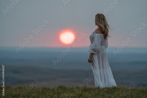 Woman standing on the top of a bluff wearing a white dress while watching the sun setting; Saskatchewan, Canada photo
