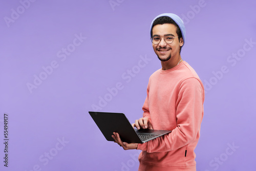 Young successful designer or freelancer in casualwear using laptop and looking at camera with smile while standing on violet background