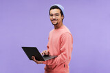 Young successful male freelancer or programmer in pink pullover, beanie hat and eyeglasses holding laptop and looking at camera