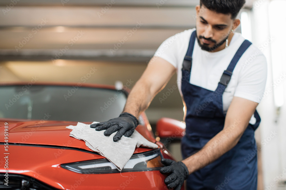 Low angle view of male professional car wash worker in black rubber gloves, holding the gray microfiber and polishing the car hood and headlight of luxury red car. Low angle view of male professional car wash worker in black rubber gloves, holding the gray microfiber and polishing the car hood and headlight of luxury red car.