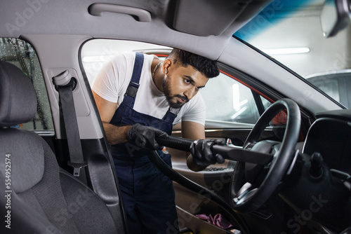 Car wash and detailing service. Bearded male worker in overalls and gloves, making chemical cleaning process with a vacuum cleaner, releasing steering wheel of the car from dust and sand.