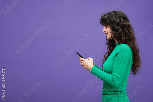 Side view of young brunette woman in green costume using mobile phone while texting or looking through online information in isolation