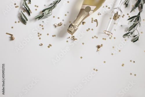 Happy New Year decorative border, web banner. Birthday, wedding party celebration. Golden champagne wine bottle, drinking glass. Olive tree branches on white table background. Star confetti. Flat lay