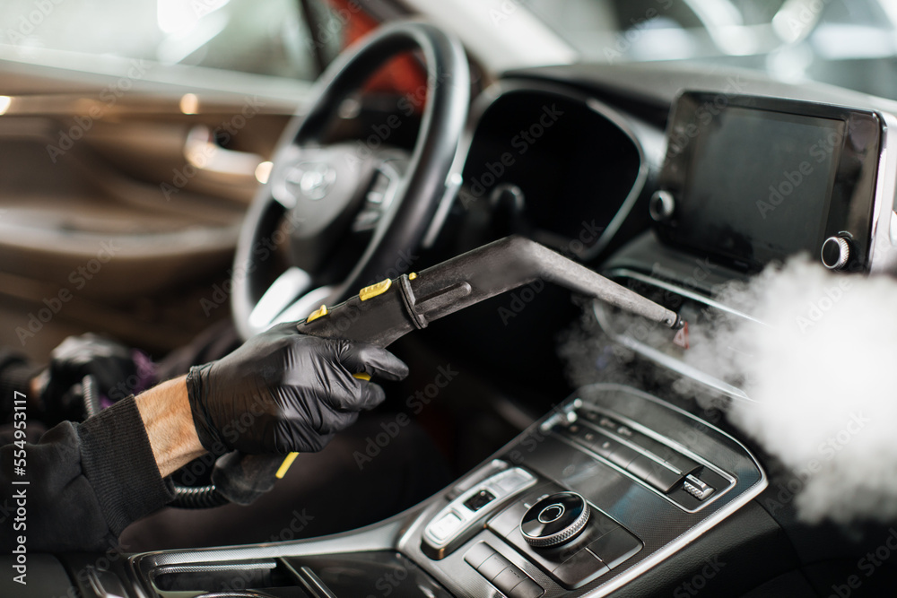 Close up of hands of man in black protective rubber gloves cleaning interior of the car with hot steam cleaner. Selective focus on guy hands. Auto cleaning service and detailing concept.