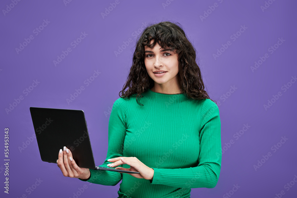 Young confident teacher or student in green pullover holding laptop and networking while standing in front of camera on violet background Young confident teacher or student in green pullover holding laptop and networking while standing in front of camera on violet background