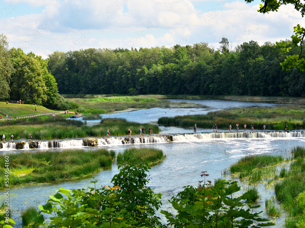 Kuldiga, Latvia - July 31, 2021: Tourists cross the river Vetna along the longest waterfall in Europe Rumba. Kuldiga, Latvia - July 31, 2021: Tourists cross the river Vetna along the longest waterfall in Europe Rumba.