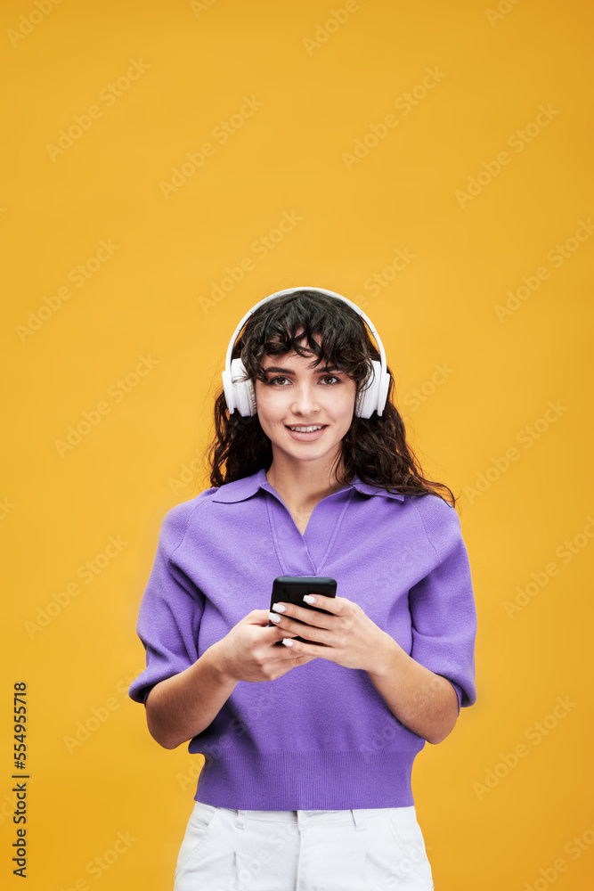 Pretty student of college in headphones looking at camera while scrolling through playlist in smartphone and choosing audio file to listen Pretty student of college in headphones looking at camera while scrolling through playlist in smartphone and choosing audio file to listen