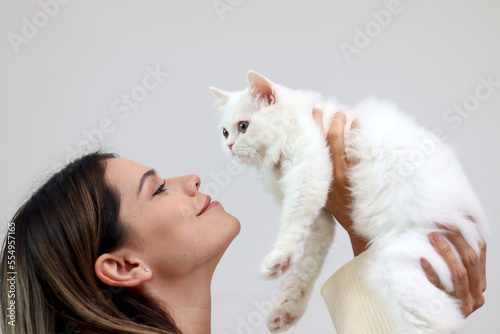 Young happy smiling woman holding cute white Persian cat and lift up her cat, try to kiss her pet, happy beautiful lady playing with fluffy pet, girl hugging her adorable long hair kitty with love