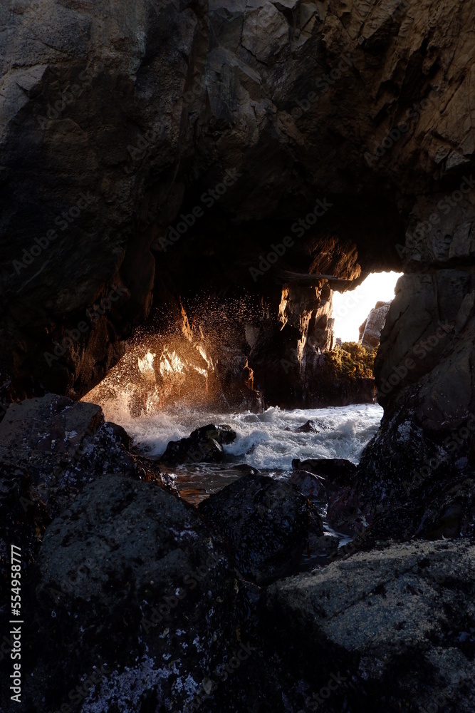 Waves rolling in from the pacific ocean and crashing into rocks at the entrance of a cave in the rays of the sun