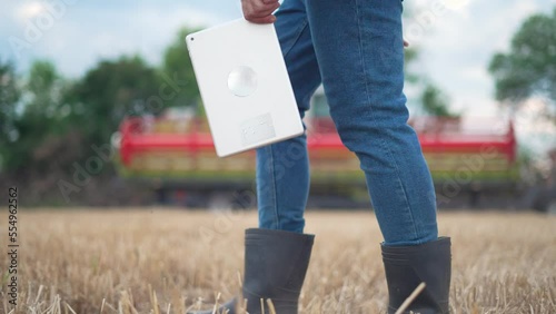 Agriculture.Farmer agronomist in rubber boots with tablet.Combine harvest control.Farmer in harvested wheat field.Agronomist work on background of combine in field.Wheat harvest on farmer plantation