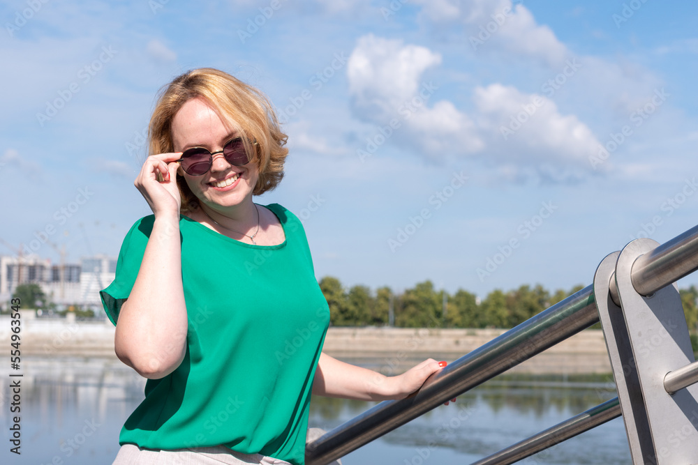 Portrait of a smiling happy girl in sunglasses, a green blouse holding glasses with her hand standing on the river bank.