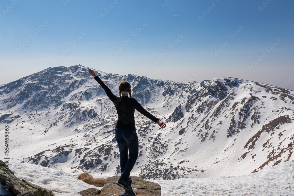 Rear view of woman on top of summit Kreiskogel spreading arms and looking at snowcapped mountain peak Zirbitzkogel, Seetal Alps, Styria, Austria, Europe. Hiking trail Central Alps in sunny winter