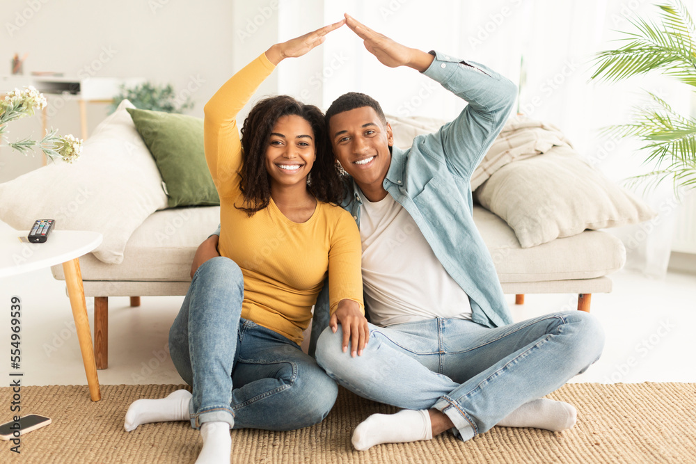 Smiling young african american wife and husband sit on floor do roof with their hands and look at camera Smiling young african american wife and husband sit on floor do roof with their hands and look at camera