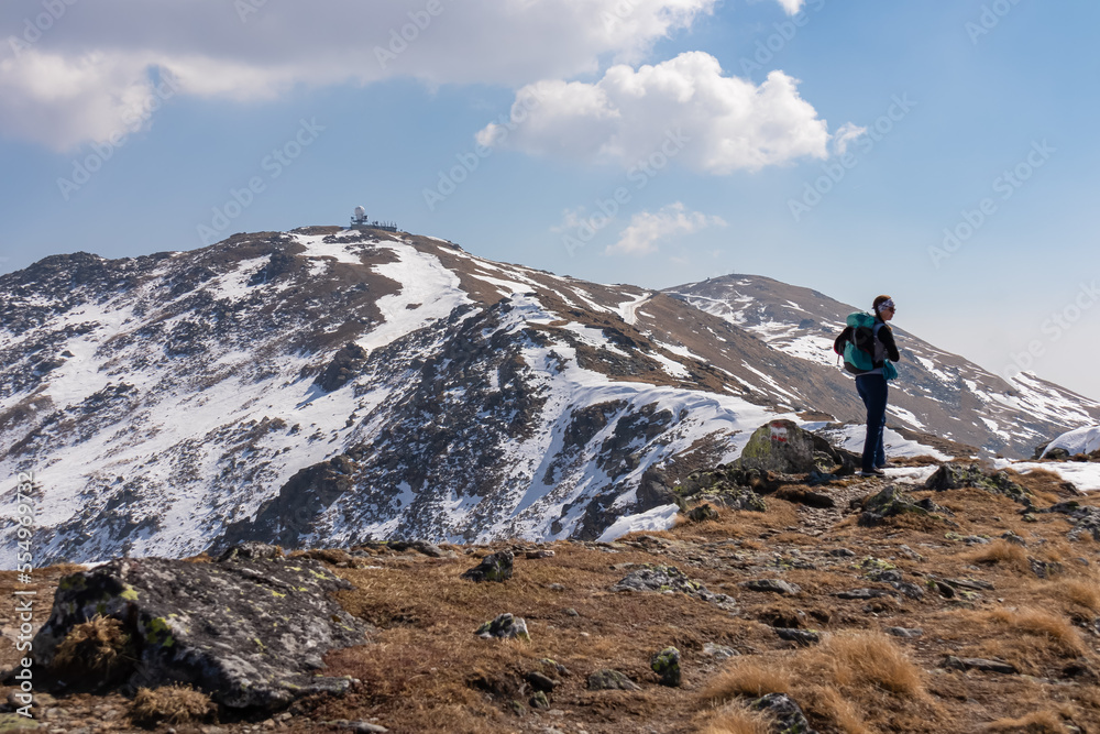 Rear view of woman with backpack hiking on snow covered hiking trail to radar station with scenic view on snow capped mountain peak Zirbitzkogel, Seetal Alps, Styria (Steiermark), Austria, Europe