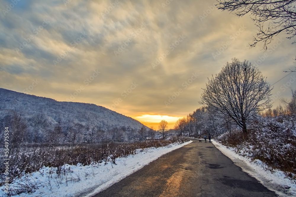 Asphalt road in winter at sunset. In the distance, small silhouettes of two pedestrians and a dog are visible. The orange sun reflects in the wet pavement. There is snow on the side