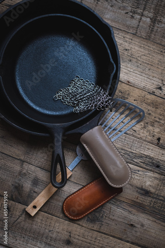 Cast iron skillets, with a metal spatula, leather handle sleeves and a chainmail scrubber on a wooden background. Cast iron cleaning and care.