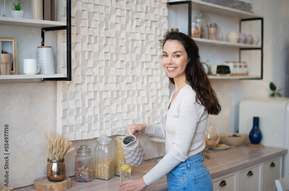 Attractive young woman in casual outfit pouring fresh water into glass in kitchen interior and smiling at camera Attractive young woman in casual outfit pouring fresh water into glass in kitchen interior and smiling at camera