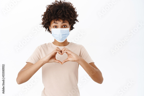 Health and healthcare. Young Black girl in medical face mask, shows heart, love sign, care about people during covid-19, white background © Cookie Studio