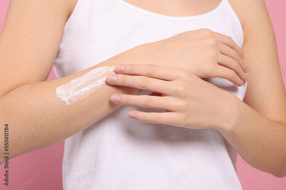 Young woman applying body cream onto arm on pink background, closeup