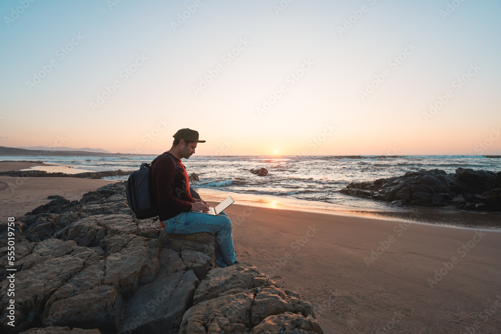 mature latino man sitting on a rock on the beach shore with his notebook working at sunset