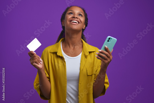 Young happy cute African American woman with mobile phone and mockup credit card in hands looking up joyfully having managed to make cool purchase in online store at good price, stands in lilac studio