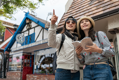 travel lifestyle and people on holiday in usa. asian Korean woman pointing at landmark in distance with her smiling friend while consulting guide in solvang California