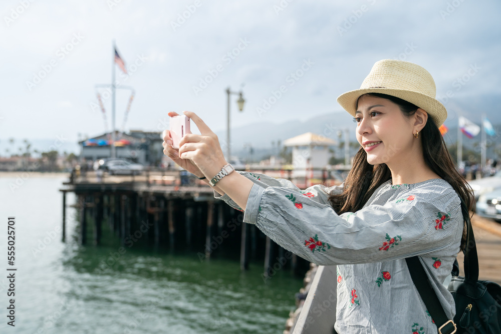 happy asian taiwanese lady traveler using smartphone to take picture of beautiful ocean view on pier while touring santa Barbara harbor in california usa