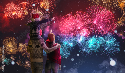 Mom and dad with their son on their shoulders in warm clothes and in a Santa hat. The family celebrates the new year looking at the fireworks outside