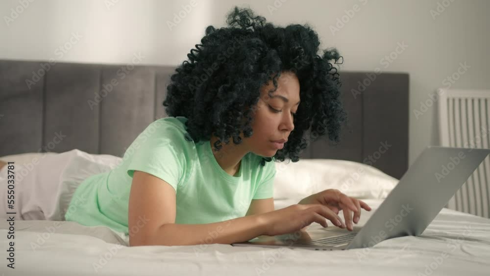 Focused African woman thinking of problem solution, watch webinar, read email, laying on bed looking at laptop learning new app, works on research project online on computer, makes remote project task Focused African woman thinking of problem solution, watch webinar, read email, laying on bed looking at laptop learning new app, works on research project online on computer, makes remote project task