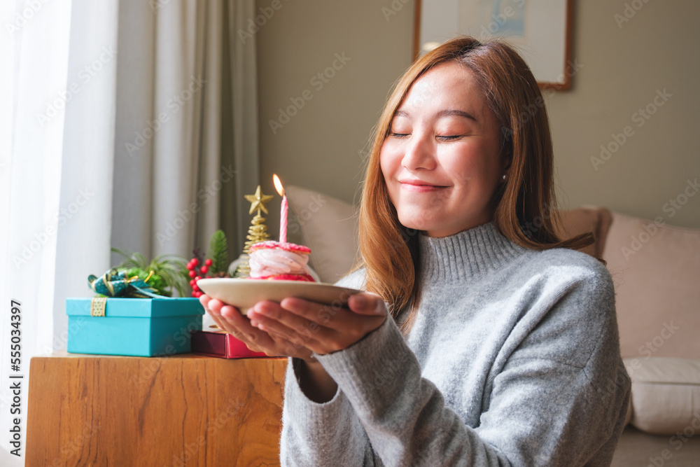 Fototapeta premium Portrait image of a young woman wishing and holding birthday cake with candle, Christmas holiday decoration at home