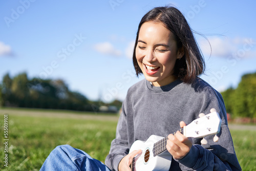 Happy people and hobbies. Smiling asian girl playing ukulele guitar and singing, sitting in park outdoors on blanket photo