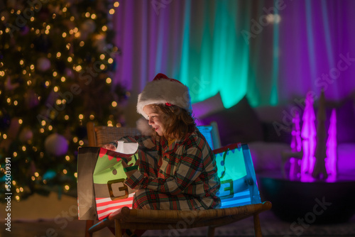 Kid with present gift with magic light. Lighting present gift bag. Happy child in pajamas near Christmas tree at home with traditional Christmas tree.