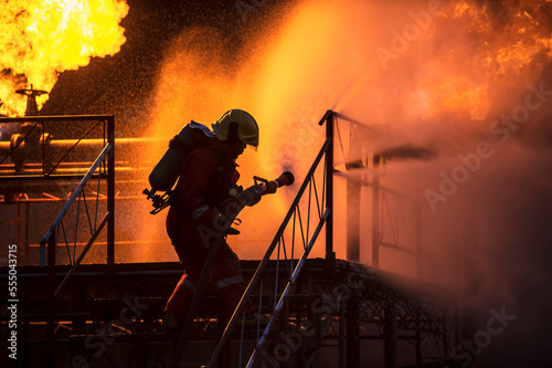 Firefighter in training with a fire hose nozzle spraying high-pressure water on a fire and wearing a fire suit for protection in a dangerous situation