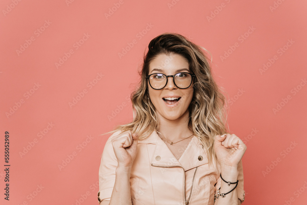 Close up portrait of a young cheerful blonde woman in glasses wearing a light pink leather jacket clenches fists, celebrates her success, over pink background. People emotions concept. Close up portrait of a young cheerful blonde woman in glasses wearing a light pink leather jacket clenches fists, celebrates her success, over pink background. People emotions concept.